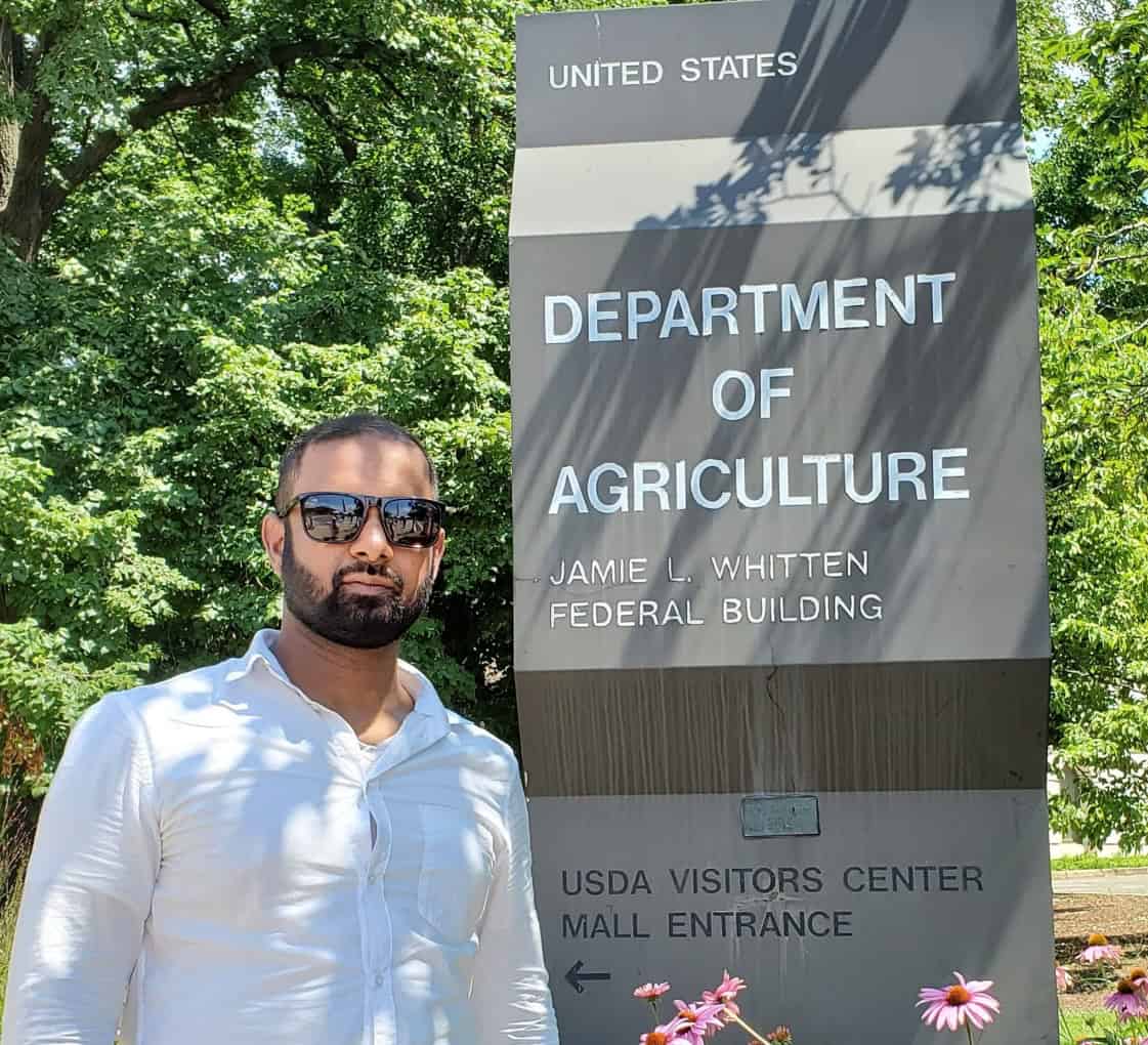 Mahfuzur 'Fuz' Rahman standing next to the Department of Agriculture sign and the Jamie L. Whitten Federal Building in front of several leafy green trees.