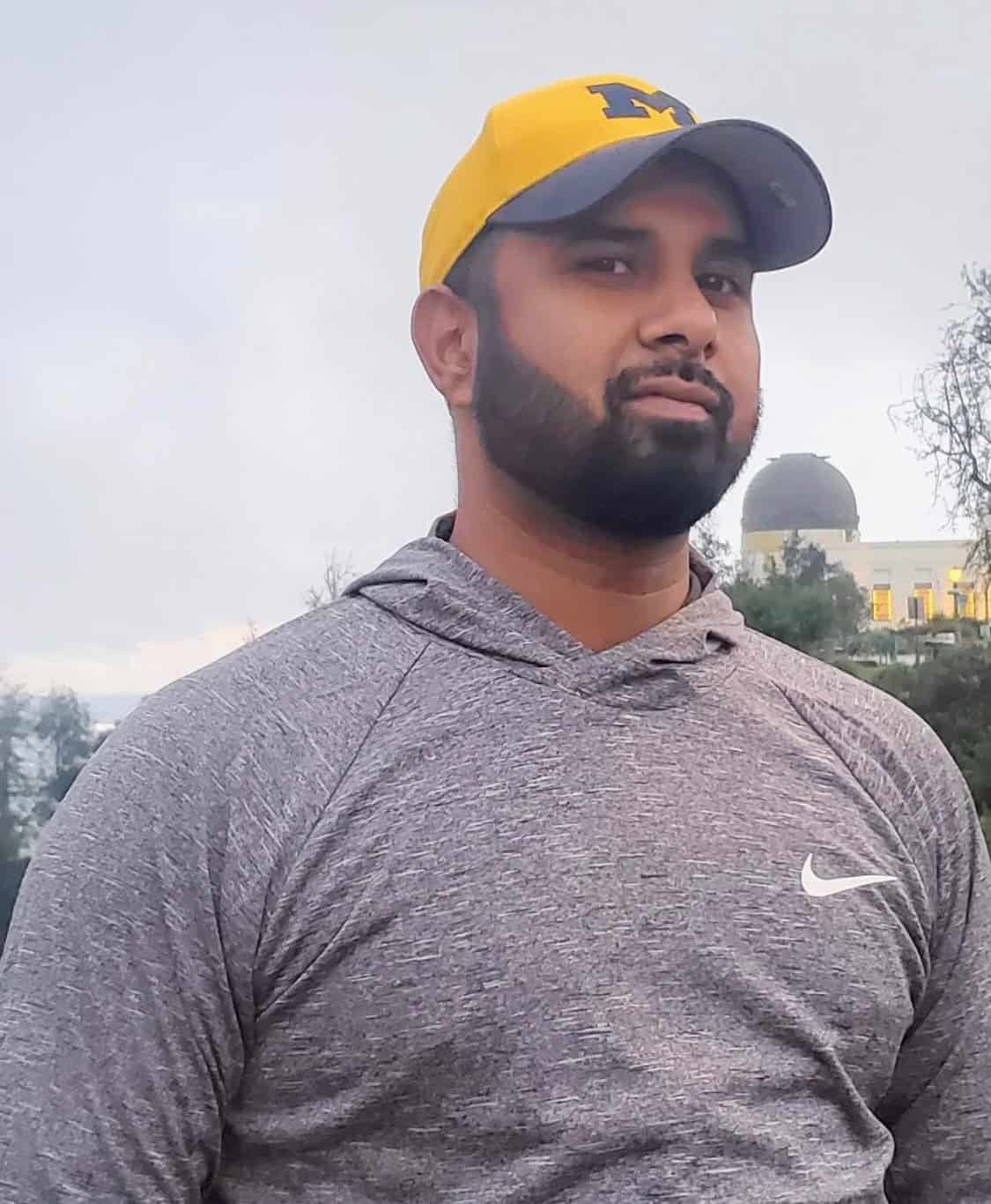 Mahfuzur 'Fuz' Rahman looks at the camera wearing a yellow baseball hat and a grey Nike hooded pullover on a cloudy day.