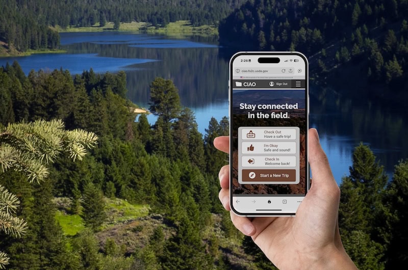 A person holds a smartphone displaying the CIAO check-in/check-out interface against a scenic forest backdrop with a lake and pine trees, representing field use in remote environments.
